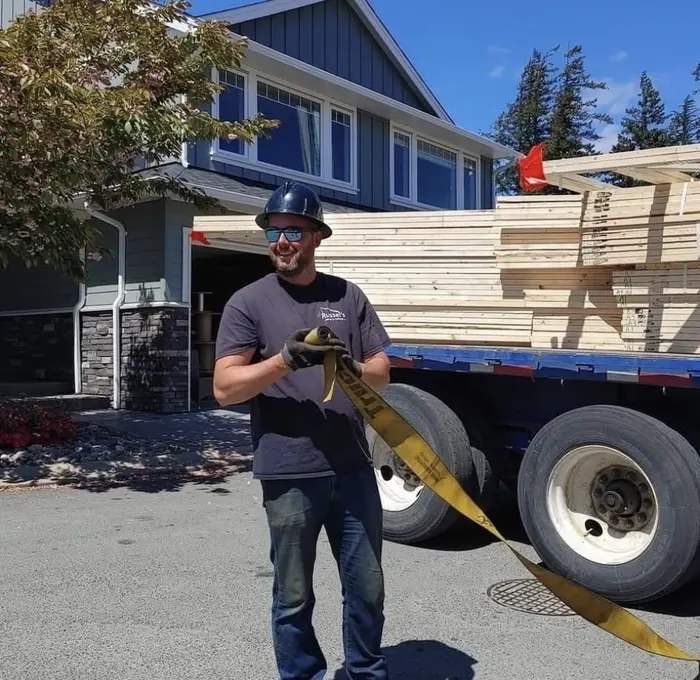 Smiling crew member in a hard hat holding a strap beside a lumber flatbed